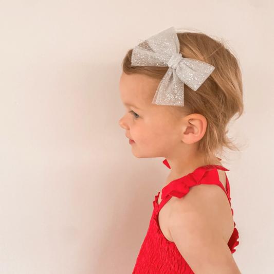 Little girl wearing a Sparkle Tulle Bow, shimmering in soft light. The bow measures approximately 10 cm wide by 9 cm tall and is available on a nylon headband for babies or right and left clips for toddlers. Perfect for adding a whimsical touch to her hairstyle for special occasions or everyday wear.